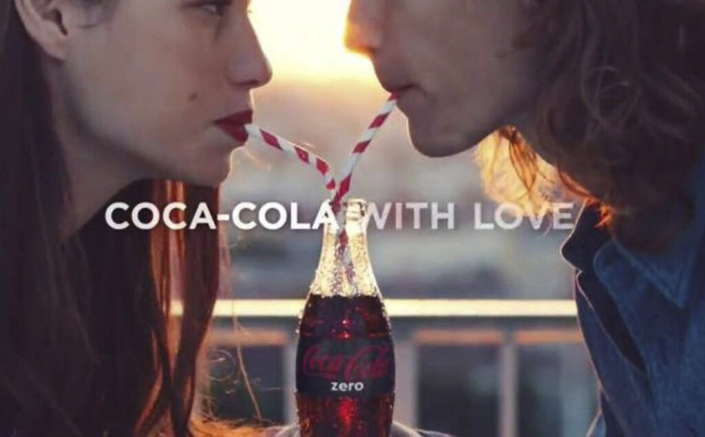 quadro de comercial, texto "coca-cola with love", imagem casal dividindo a mesma garrafa de coca-cola zero, cada um com um canudo, ela olha em seus olhos.