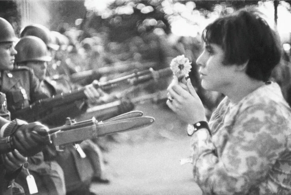 Foto em preto e branco. à esquerda, soldados apontam armas. À direita, mulher segura um flor, voltada para eles.