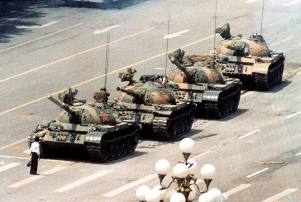 Foto colorida. Em uma rua, fila de quatro tanques de guerra e um homem sozinho voltado para eles.