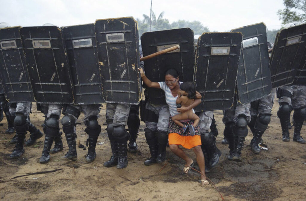 Foto colorida. Nove militares lado a lado cobertos pelos escudos que seguram empurram mulher que segura uma criança pequena no colo e tem expressão de medo.